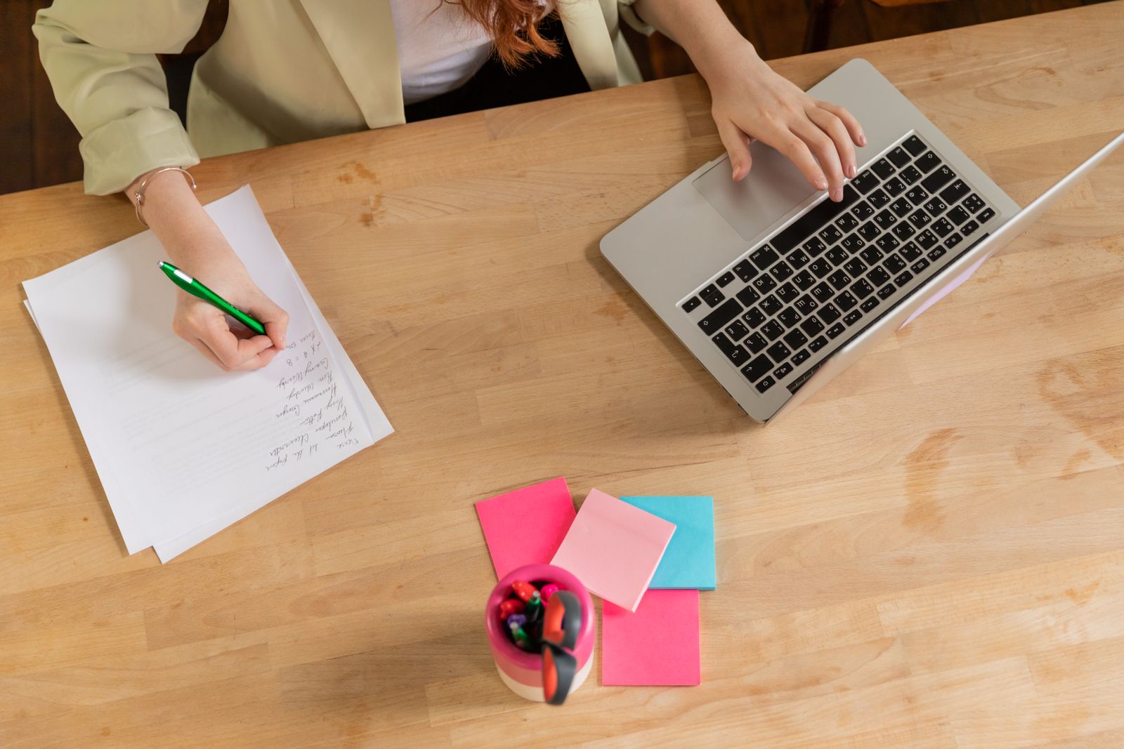 Overhead view of a person writing a handwritten shortlist on paper next to an open laptop on a wooden desk