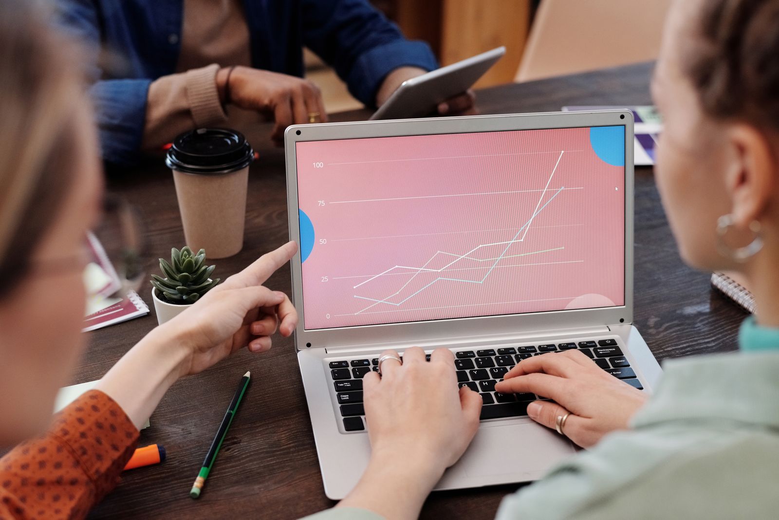 One colleague points at a line chart on a laptop screen while another types, during a meeting at a wooden table