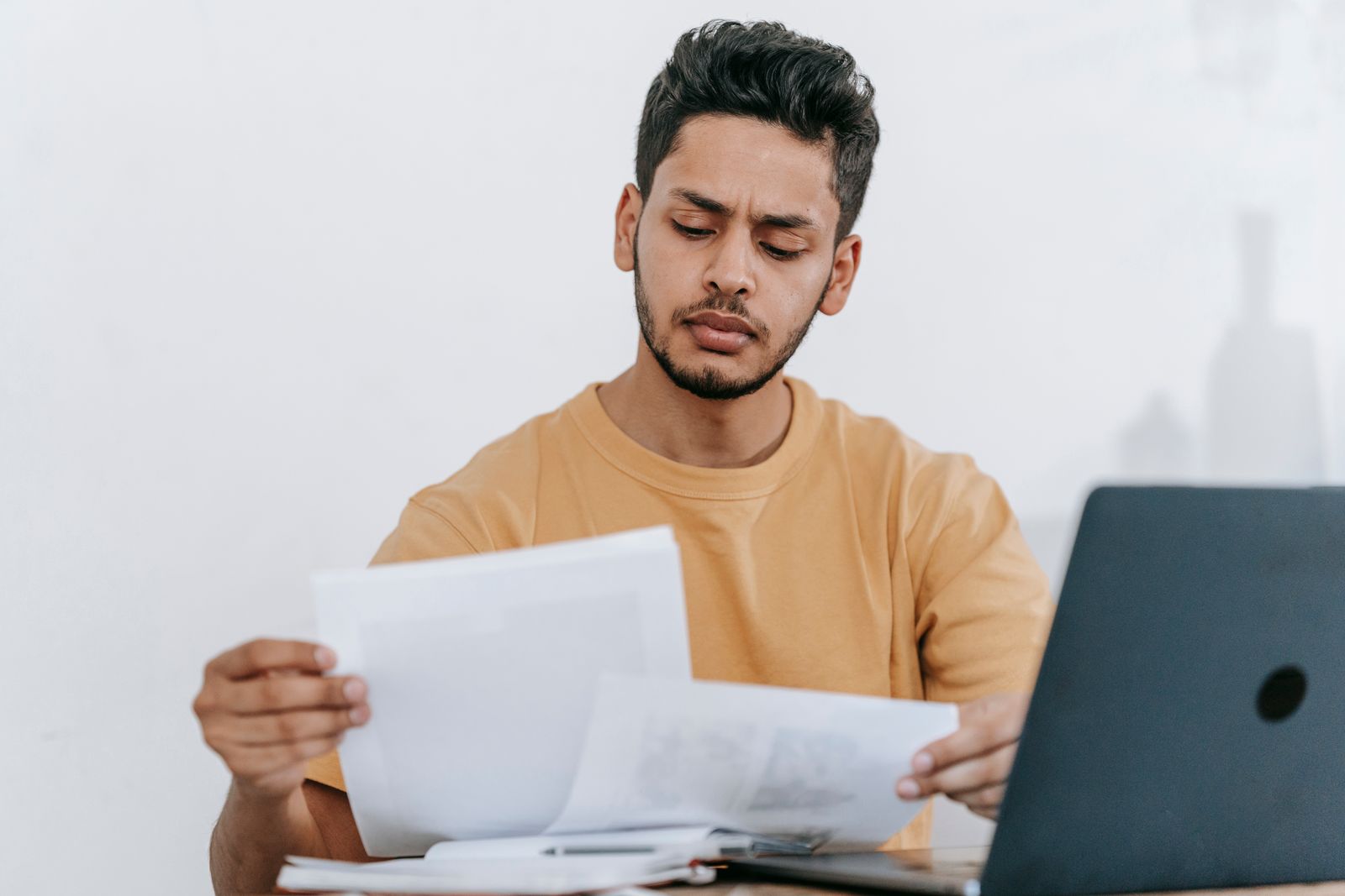 A young man in a mustard T-shirt frowns as he compares two printed pages at a desk beside an open laptop