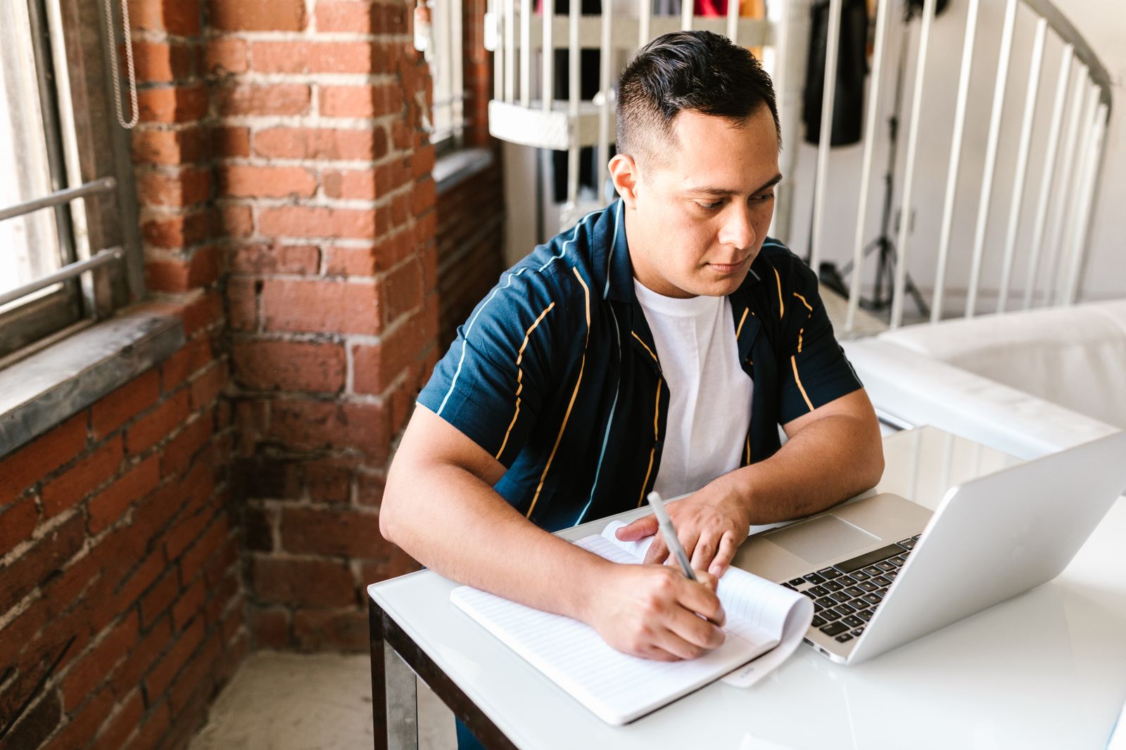 Man writing notes in a lined notebook beside an open laptop at a small table by a window