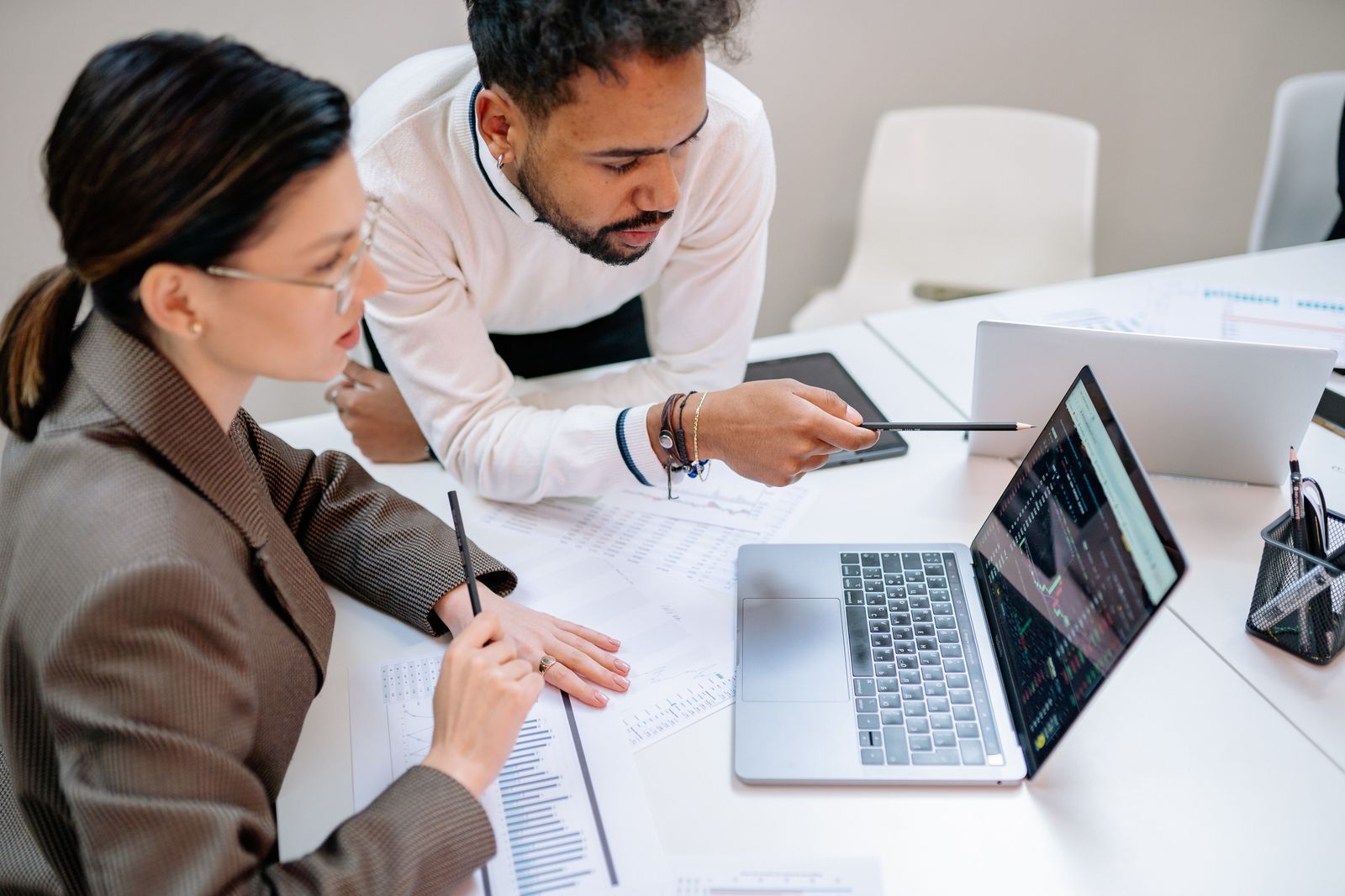 Two colleagues at a white desk pointing at a laptop screen and printed charts during a data review