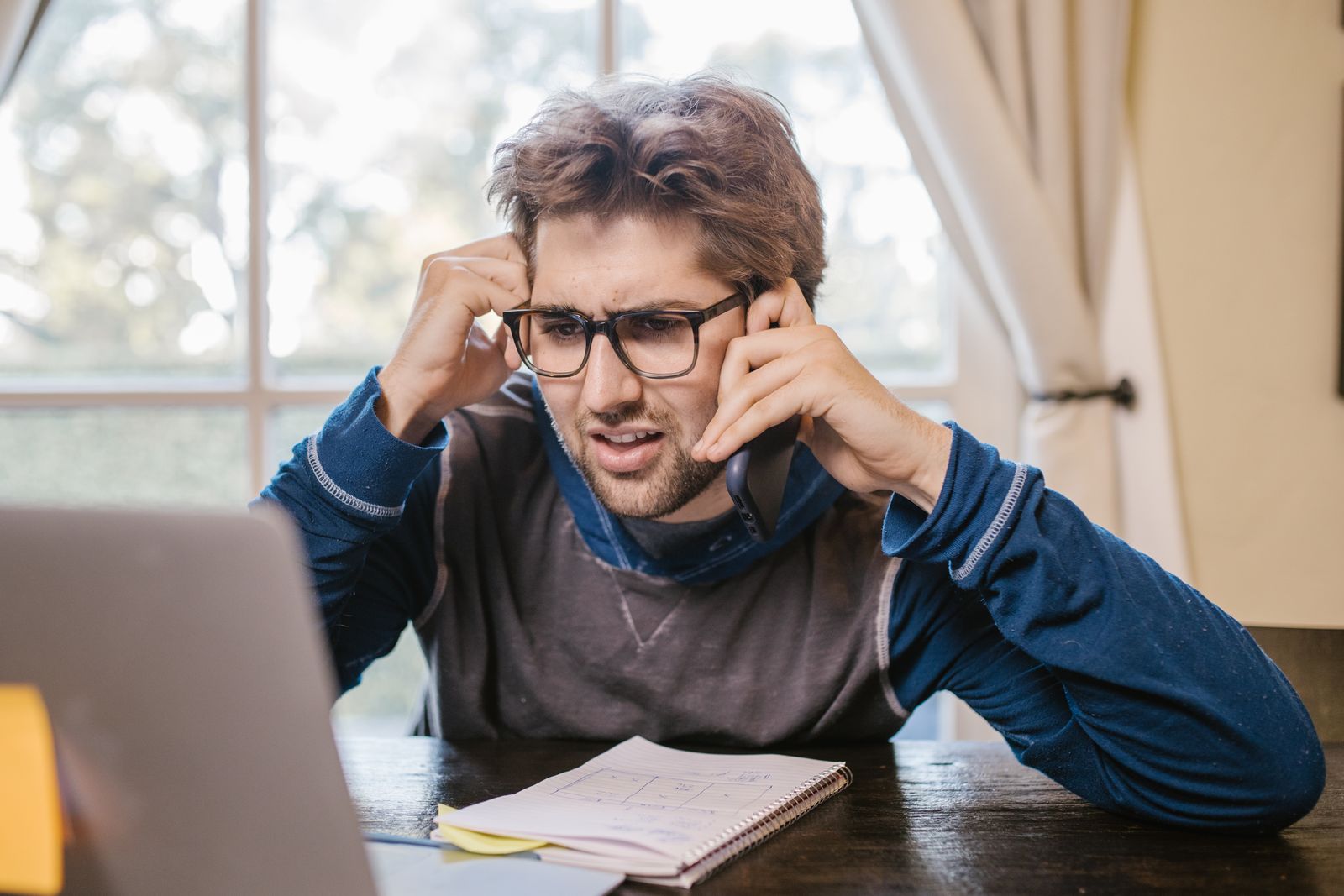 A young man in glasses holds a phone to his ear and frowns at his laptop, a notebook open on the table in front of him