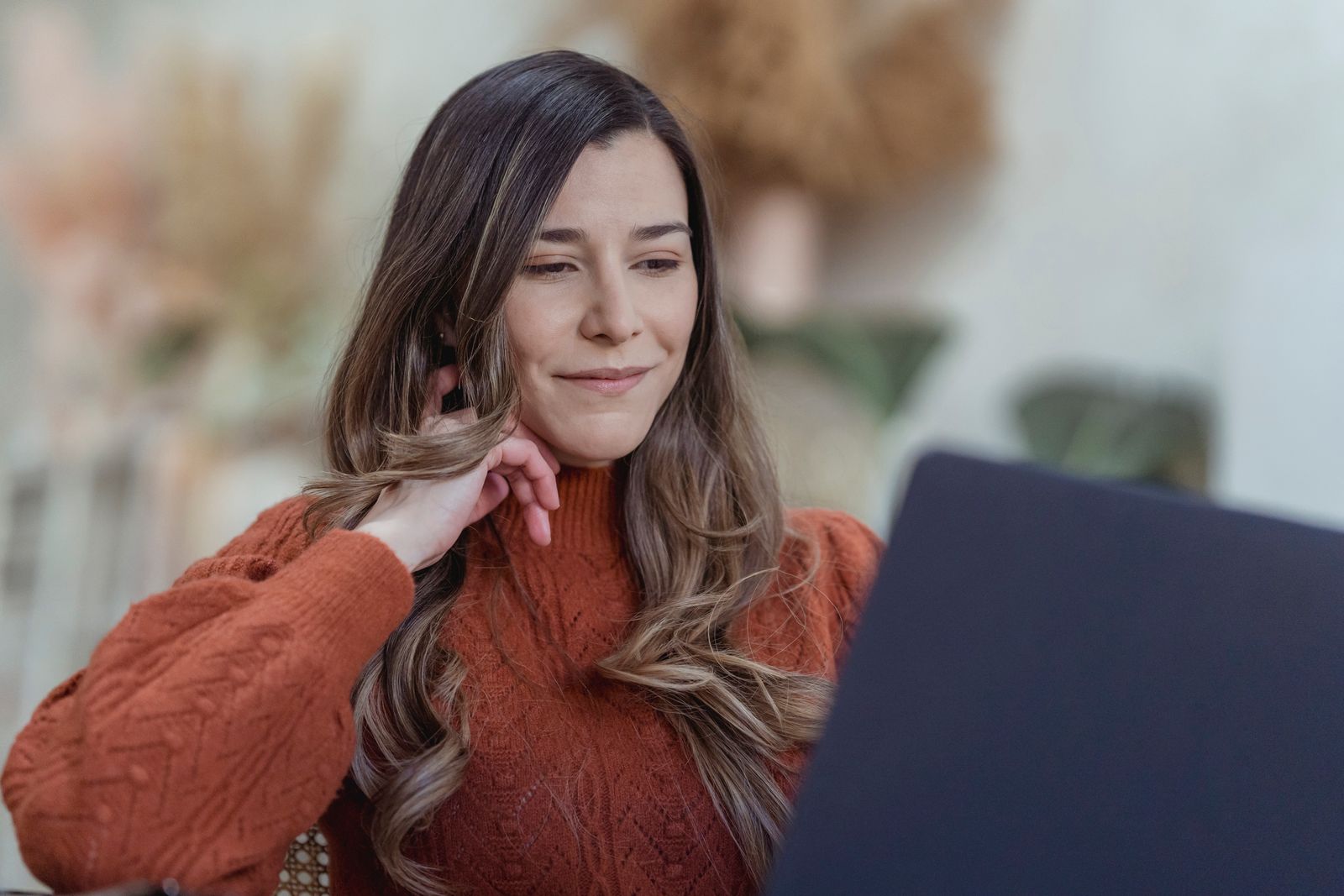 A woman in an orange sweater sits at a laptop at home, reading the screen with a thoughtful half-smile