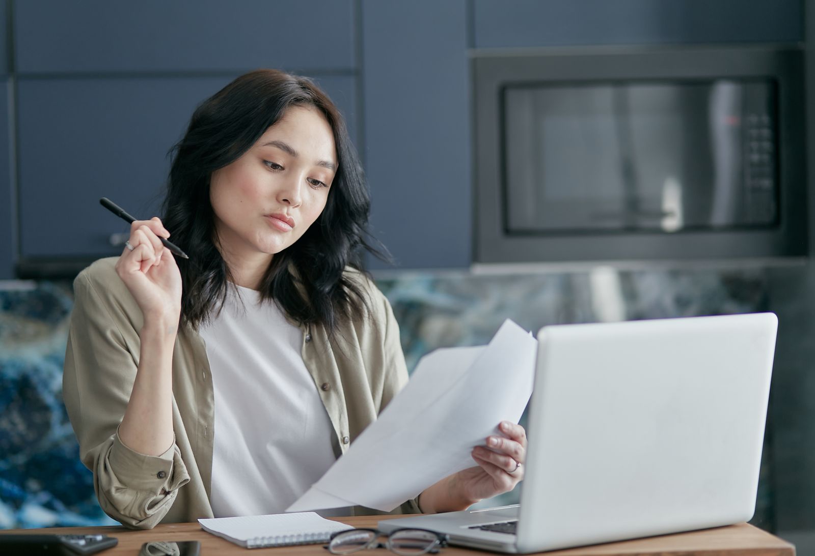 A woman at a kitchen table holds printed pages and a pen, reading them with a doubtful look beside an open laptop
