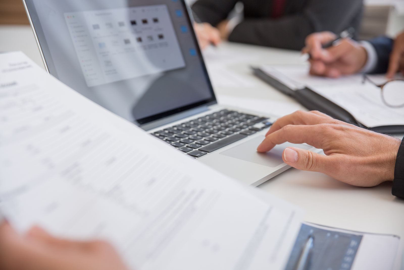 A printed document held in the foreground while a hand rests on the trackpad of an open laptop on a white office desk