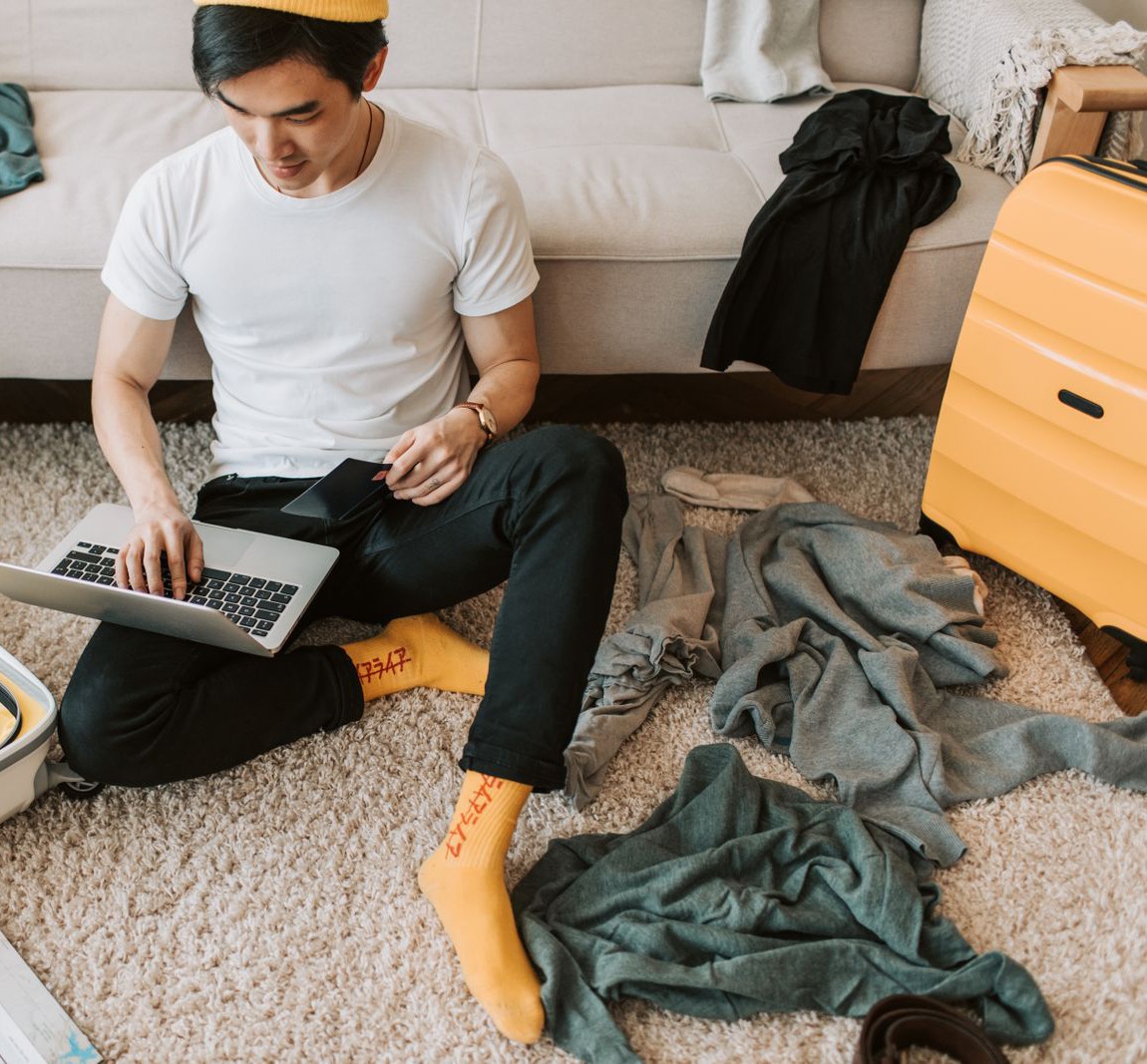 A young man sits on a living room rug with a laptop on his knees and a travel document in his hand, next to a yellow suitcase and clothes laid out for packing