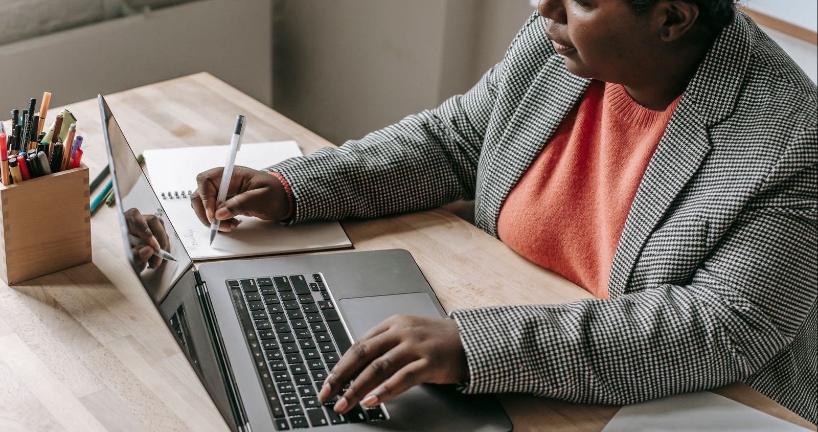 A woman in a houndstooth blazer types on a laptop with one hand and writes in a notebook with the other at a wooden desk with a pot of pens