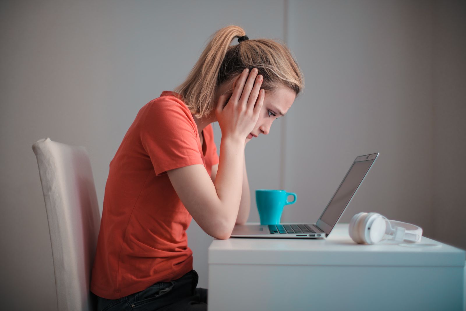 A young woman in an orange T-shirt holds her head in her hands as she stares at an open laptop beside a coffee mug and headphones
