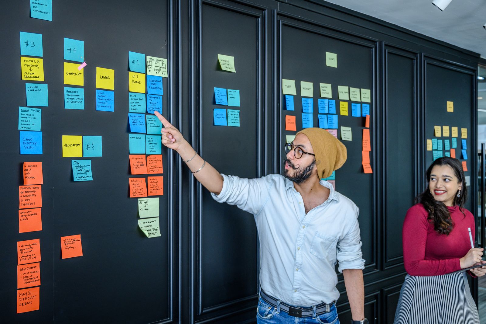 A man points at a numbered column of sticky notes on a dark wall during a planning session while a colleague with a tablet looks on