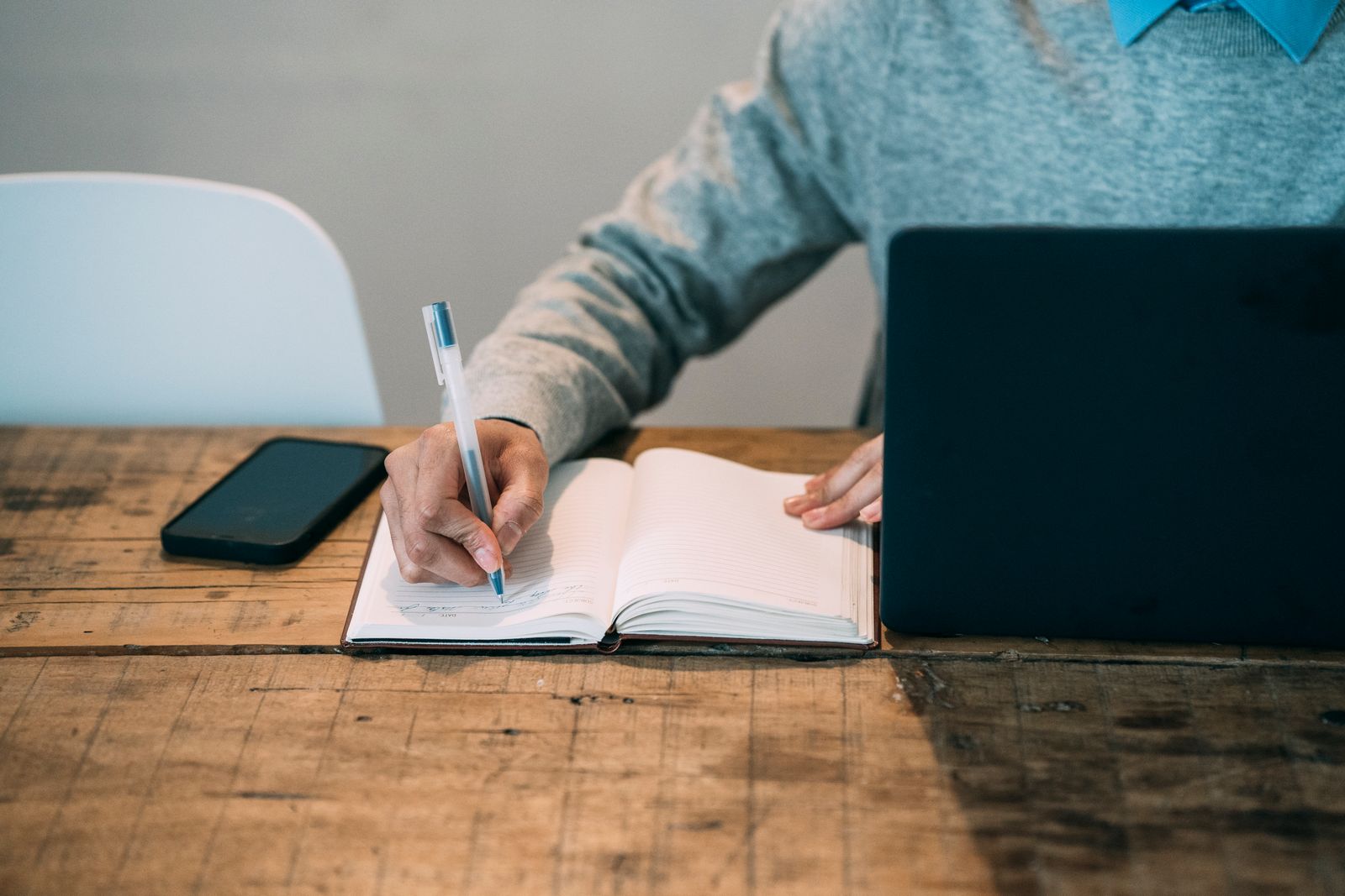 A person in a grey sweater writes notes in an open notebook beside a laptop and a phone on a worn wooden table